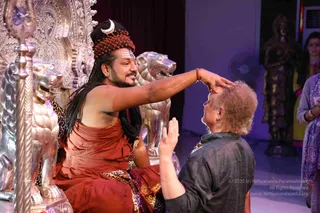A seated man in a rust-red and saffron-toned robe, with long dark hair, a rudraksha bead crown and a crescent ornament at the top of his head, and a tilak on his forehead, extends his right hand to touch the forehead of a kneeling older man in a dark grey kurta. Behind him stand large silver lion figures and an ornate silver throne with carved floral panels, with a tall silver standing figure and a silver staff in the foreground. Swamiji (probable) is the seated figure in the rudraksha crown and red robe, shown blessing the kneeling devotee on a stage setting. Two women in colourful dress stand at the right edge.