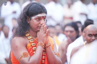 A person with long, swept-back dark hair stands in front of a crowd of people dressed in white, with hands pressed together in namaste near the chest. He wears an orange-saffron shawl draped over the shoulder and bare arm, a multi-strand gold and rudraksha-style bead necklace with large golden beads, and coral-red beaded armlets and bracelets. A white sacred ash mark with a red bindi is on the forehead. Swamiji (probable): the long hair, saffron shawl, ornate beaded gold necklace and armlets match Swamiji's distinctive presentation, and the page indicates he attends this event. Behind him, devotees in white clothing stand in a blurred gathering in an outdoor temple setting.