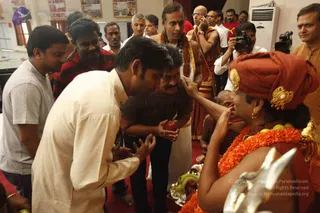 A photograph of devotees gathered in a hall with red carpet during a ritual encounter. In the foreground, a man in a cream kurta leans forward with palms open toward a woman in a red-and-gold head wrap and orange marigold garland, who touches a man's forehead; a man in a dark t-shirt holds a red fruit or offering. Behind them, a bald man in a white dhoti and shawl holds a phone and a man with a beaded necklace stands nearby. A cameraman films and a tray with bananas and fruit sits in front. Wall posters and an air conditioner are visible. a person's presence is not clearly established from appearance.