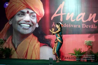 A young female dancer in a teal and gold traditional costume performs on a green-carpeted stage in front of a large backdrop. The backdrop shows a huge printed portrait of a smiling man with an orange turban, red bindi, and orange shawl, with the word "Anan" in pink script and "Ānandeśvara Devālaya" below. Printed text labels the backdrop as a person. The dancer wears a white flower in her hair, bangles, and ankle bells, and holds her hands in an expressive mudra. Potted plants flank the stage.