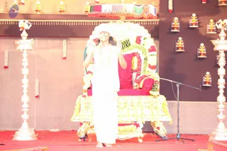 A barefoot person in a white dhoti-style garment and white shawl stands on a stage in front of an ornately decorated golden throne draped in red cushions and garlands of white and yellow flowers. The person has a white cloth on the head, smiles broadly and raises both hands near the face. A microphone stand stands to the right, and tall silver oil lamps flank the throne. Small lamps are mounted on the dark wall behind, with a striped pink and green canopy above. Red carpet covers the stage floor. The image is faded and washed out.
