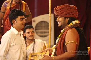 A photograph at an indoor satsang setting with a red drapery backdrop. On the right, a man with a rust-orange turban, long swept-back hair, a rudraksha mala and a flower garland stands bare-shouldered in an orange-red shawl with a gold armlet, smiling broadly. Swamiji (probable) by appearance cues: turban, long hair, saffron-red clothing and rudraksha mala, with the role of a teacher receiving devotion. Facing him on the left, a young man in a white shirt smiles and holds a framed gold-bordered card or photo. Behind them, a priest in orange-and-white robes with a rudraksha mala and a second man in white with a red tilak stand near a white fan and a gold pole.