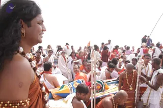 A photograph taken aboard a boat on the Ganga during a boating visit at Varanasi. In the left foreground a smiling person with long dark hair wears an orange-saffron garment, a large necklace of rudraksha beads and cowrie-like shells, and a dotted white mark on the forehead; the page's a person reference gives an appearance-based candidate, but the cues are limited, so this is marked unsure. Behind them a crowd of devotees in white and orange-saffron robes, some with red shawls, sits on the boat and in other boats. Near the centre a bald man in orange robes and a gold pendant chain holds a long silver staff with a trident-like finial, and a person in white kneels in front of a colourful patterned cushion. A ritual plate is visible among the devotees, and the sky is bright white over the river.