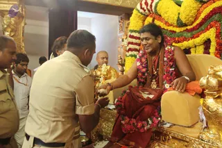 A seated man in a red-maroon dhoti and a bare chest wearing layered rudraksha malas and a gold pendant medallion sits on a golden throne decorated with floral garlands in yellow, orange, red and pink. A uniformed officer in khaki shirt and trousers stands bending forward, extending his hand toward the seated man, who reaches out and receives or blesses him. Behind them a bald priest in white dhoti and a golden deity murti or ritual brass pot stand in a gold-ornamented shrine room, with a gold lion figure in the right corner. Several other onlookers in white shirts stand at the left edge. Overall a temple-style ceremonial darshan setting with gold decor and flower garlands.