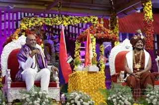 Two men sit on ornate silver-and-red thrones on a flower-decorated stage with marigold and rose garlands overhead. On the left, a man in white trousers and a purple shawl with a rudraksha mala holds a microphone and smiles. On the right, a man in a rust-brown robe and matching turban wears layered gold necklaces and sits beside a brown-robed attendant with a crown-like ornament. Between them stands a gold rose-covered table with a silver vase of white flowers, flanked by red and saffron flags, one showing a Mauritius flag.