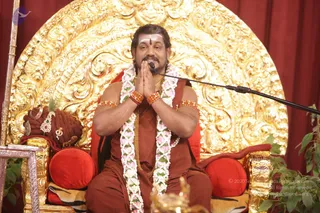 A man seated on a red-cushioned golden throne with an ornate gilded back in front of red curtains, palms pressed together in namaste. He wears a rust-orange robe, white and pink flower garlands, orange rudraksha bracelets on his wrists and upper arms, a gold chain, and a white tilak with a red dot on his forehead. A microphone on a stand is in front of him. Beside the throne is a brown ceremonial seat with gold jewelled ornaments and a small crown-like item. Swamiji (probable): long swept-back hair, orange-red robe, rudraksha bracelets and gold necklace, seated on a throne during a discourse.