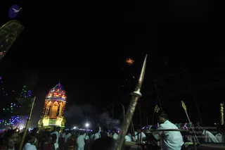 Night procession with a brightly illuminated ratha chariot topped by a tiered tower lit in yellow, red and green lights, moving through a dark crowd. Many people in white clothing walk and raise hands, while a man in a white shirt in the right foreground holds up a long ceremonial trident (trishul) and others carry tall brass-tipped staffs. A single firework bursts in the night sky above. Strings of coloured lights and a flag edge the left side.