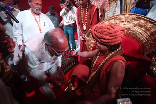 A photograph of a ceremonial gathering on a red-carpeted stage. In the centre, a figure seated on an ornate gold-and-red throne wears a saffron-orange turban, a saffron robe, and layered gold-bead necklaces over a bare shoulder, and is smiling while reaching out. Before them, a balding man in white kurta with a sash bends forward and reaches toward the seated figure's hand, appearing to pay respects. Behind them, a man with a grey beard in white kurta and an orange lanyard stands, and a person in a red-brown robe with gold malas stands at the back. A man with a camera and others in the background observe, with a microphone at the left edge. The seated figure's cues of turban, ornate gold necklaces and saffron attire make this a probable appearance of Swamiji, seated in person on the throne.