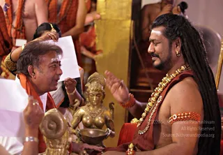 A photograph of two men seated side by side in a temple-like hall with a golden pillar behind them. On the right, a bare-chested man with long swept-back dreadlocks, a white tilak on his forehead, a dense gold and bead necklace, a red-orange rudraksha bracelet and a coral armband sits with eyes closed and one hand raised in gesture. Next to him stands a golden statue of a female deity holding a bowl. On the left, a man in an orange shawl and gold bangle gestures while speaking, and a hand holds a white paper. Behind, other people in orange clothing are visible. The most prominent figure has long swept-back hair, ornate gold necklace and an orange wrap, consistent with the appearance-based a person label, seated in ritual dialogue.
