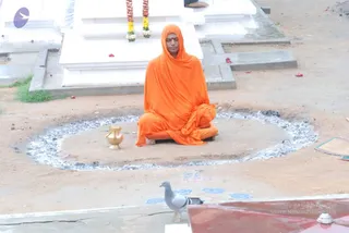 A photograph of a person seated cross-legged on a bare earthen platform inside a circle of white ash, in a courtyard beside white stone structures with garlands of yellow and orange flowers hanging from them. The seated figure wears an orange saffron shawl draped over the head and body and matching orange cloth wrapped around the legs, with a red tilak on the forehead, eyes closed in meditation. A brass kalasha/pot sits to the left of the circle. A grey pigeon stands at the lower centre near a red-painted ledge, and faint blue and white painted markings lie on the ground. Scattered flower petals lie around the platform. The figure appears to be a sannyasi in saffron robes, though identity cannot be confirmed from appearance alone.