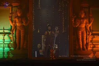 A photograph of a dimly lit stage tableau set between two tall, ornately carved golden-orange statues of multi-armed deities with raised arms, flanking a central niche strung with small fairy lights and hanging bells. In the centre stands a single figure in a dark maroon-brown robe with a gold necklace, holding a silver trident upright beside a golden kalasha-like altar piece. A zebra-striped seat sits behind him. Teal and amber stage lighting washes over the carved pillars. No other people are visible.