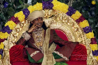 A photograph of a man seated on an ornate gold throne with a red velvet cushion, framed by garlands of yellow marigolds and purple flowers against green foliage. He wears a beige turban with a red bindi, a maroon and gold embroidered sherwani, a heavy gold necklace and pendant, and a cream shawl draped over his lap. He rests one hand on his cheek and smiles to the side. The setting appears to be a ceremonial seat during a public event. Swamiji (probable) appearance cues: turban, ornate ceremonial clothing and gold necklaces on a throne.