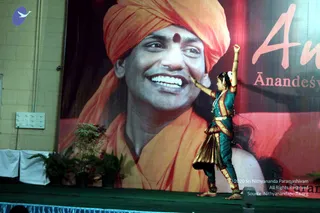 A female classical dancer in a teal and gold costume performs on a green-skirted stage, one arm raised high and the other extended toward a large banner. The banner shows a smiling man in an orange turban with a red bindi and orange robes, with partial red-and-white lettering reading "An" and "Ānandeśv" beside him. Potted plants with ferns sit at the stage edge, and a blue bird logo appears at the upper left of the wall. The photo is cropped and shows only the dancer as a physical person.