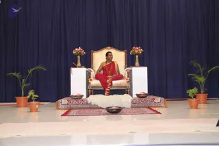 A seated man in red robes sits on an ornate gilded throne-style armchair with a cream cushion on a stage, a deity-like ceremonial setting. He wears a red shawl over a red dhoti-style garment, a long rudraksha-style mala necklace, and gold trim on his cloth. Flanking the throne are two white pedestals with flower vases of pink blooms, potted palms in terracotta pots, a red and gold patterned rug, a white fur-like cushion, and offering bowls with petals on the floor. The backdrop is a deep navy blue curtain. Only one person is visible. a person: the seated man has cues of a red and gold ceremonial robe, a rudraksha mala and a throne-like seat, consistent with the page's discourse event.