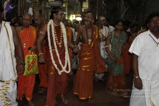 A procession inside a temple corridor with carved pillars and a lit lamp in the background. At centre, a man in a rust-red dhoti and a long white flower garland walks hand in hand with an elderly bald man wearing a bare upper body, orange-and-gold striped cloth, a rudraksha mala and a large gold necklace. Behind them, men in orange robes and white dhotis, one holding a green shopping bag, and a woman in a teal sari with gold border walk in the crowd. A man in white on the right wears a red-beaded mala. The scene reads as a temple procession with devotees.