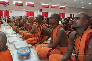 Rows of shaven-headed sannyasis seated cross-legged on silver-grey floor mats in a bright hall with red curtains and white ceiling lights. They wear saffron-orange robes with red borders, rudraksha mala necklaces and sacred ash markings on their foreheads. Small brass lamps and a clear plastic tub of offerings sit before them. Behind the rows, devotees in white and other saffron-clad attendees are visible, and a few people stand at the far left.