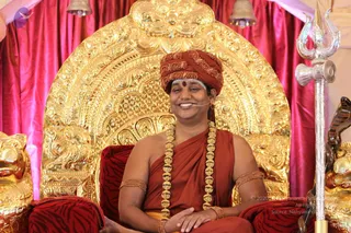 A man seated on an ornate gold throne with a carved backrest, smiling, wearing a red-orange turban with gold embroidery, a red bindi, orange-red robes and a long gold bead necklace. Gold bangles are on his upper arms and wrists, and a small microphone is at his face. A silver trident stands at right with a silver cobra shape beside it, and red velvet drapes and a red-and-black striped cushion frame the scene. Swamiji (probable) appears as the seated speaker on the throne, identified by the ceremonial gold necklace, saffron-orange robes and turban cues.