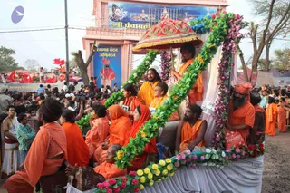 Outdoor procession at a Kumbh Mela-style gathering. A flower-decorated chariot with a white cloth canopy and a red-and-gold umbrella carries several saffron-robed sadhus and sannyasis seated on white-draped seats. A man with long hair in an orange robe stands centre-left among them, and a bearded man in orange holds the umbrella. Garlands of green leaves, purple and yellow marigolds and red flowers frame the chariot. Crowds in white and orange line the street, with red flags behind. A pink gateway above shows a painted deity mural and the text Shri Panchayati. Other saffron-clad figures walk beside the chariot.