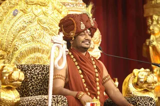 A photograph of a seated man in a rust-red turban with a jewelled ornament and white feather, a red-orange shawl and a long gold bead necklace, with gold armbands and a red forehead mark. He sits on an ornate gold throne with a leopard-print cushion and gold lion-head armrests, a microphone on a stand to his right and a red curtain behind him, indicating a satsang setting.