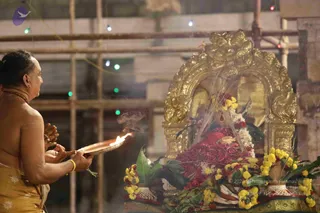 A bare-chested priest in a golden-yellow dhoti holds a brass aarti plate with a lit flame toward a deity murti decorated with marigold garlands and red flower petals. The murti sits within an ornate gold arched frame, with yellow marigold garlands arranged at its base and green leaves scattered around. Scaffolding and strings of small colourful lights are visible in the background, and smoke rises from the lamp. The priest wears a yellow sacred thread and a gold bangle and his hands are raised over the flame in an aarti offering.