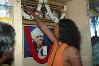 A man with long swept-back dark hair, wearing an orange saffron wrap and a long rudraksha mala with bead necklaces, stands at right reaching up with one raised arm to touch a framed gold-bordered picture on a wall. The framed picture is garlanded with white flower strands and red flowers, and beneath it a second framed portrait labelled with a printed name shows a smiling man in a white turban and dark embroidered jacket, with a red garland draped around it. At left, a partially visible man with grey hair looks toward the wall. Shelves with yellow packaging sit at left. The bare-chested saffron-clad man has distinctive long hair and rudraksha beads, consistent with a probable Swamiji appearance, while the portrait is a depiction. Total people visible: two physical people.