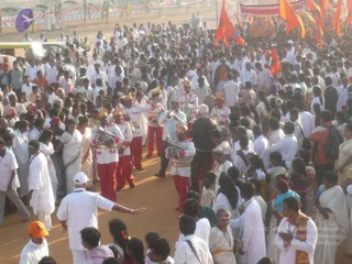 A dense outdoor crowd gathers on a dusty orange-red path during a procession. Brass band musicians in white jackets with gold trim and red trousers play tubas, trumpets and drums in the centre. Men and women in white dhotis, saris and kurtas fill the scene, with saffron flags and a decorated float or chariot rising at the top right. A man in a white cap directs with an outstretched arm at lower left. Garlands, a bright red bag and orange-red headwear appear among the people, and a bunting-decorated arch runs along the top edge.