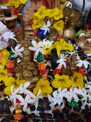 Close-up photo of a home shrine arranged with brass deity murti decorated with white jasmine flowers, yellow trumpet flowers, and red, orange and green satin garlands with gold beads. A brass Ganesha figure sits at the front-left, with a small brass murti of a seated sage or deity to the right and a taller figure with yellow flowers and a blue cloth behind. A leopard-print cloth covers the surface, with brass vessels, a bell-like brass object and small decorative items in the background. No people are visible.