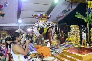 A photograph of a temple-style ritual in a decorated tent with a tiled floor. In the centre, a man in white dhoti stands with folded hands before a golden throne-like seat covered in flower garlands, while a bare-chested man in an orange cloth kneels with a towel and a silver plate beside him. A woman in a red sari stands to the right, and a photographer in a white dhoti with a camera occupies the lower left. A heart-shaped ornamental backdrop, a golden deity murti decorated with garlands, a banana plant, and an ornate oil lamp stand are visible. Hanging lamps and marigold strings decorate the ceiling.