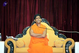 A man seated on an ornate golden throne-style armchair with black scrollwork armrests and round gold cushions, in front of a deep maroon velvet curtain. He wears a sleeveless saffron-orange robe, has a red tilak mark on his forehead, and holds his arms folded across his chest while speaking into a microphone on a stand. Bottles of water and papers sit on a small table at left. A purple logo appears in the top-left corner. Swamiji (probable): long swept-back dark hair and saffron robe on a throne, matching the page's claim of his presence at this event. Single seated speaker, no other people visible.