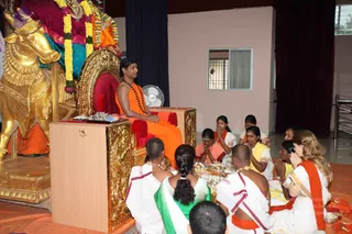 A man seated on a gold-and-red throne in a hall, wearing an orange robe, rudraksha mala beads and bare-chested with a shawl draped across his lap, hands folded. Before him kneel devotees: a shaved-headed man in white dhoti with orange-banded cloth, a woman in a green sari, women in saffron and yellow saris with hands joined, and a woman in an orange top at right, some holding brass plates of offerings. Behind the throne stands a large golden bull (Nandi) decorated with yellow and orange garlands. A white table fan and a barred window are visible. a person is identified here by the orange robe, rudraksha mala and bare-chested presentation on the throne, distinct from the white-clad priests.