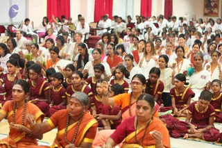 A large seated congregation fills a bright hall with red curtains and white walls, with a framed painting at upper right. In the foreground, several women wear orange and yellow saris with rudraksha malas and bindi-style forehead marks, some with palms raised in gesture. Behind them, rows of girls and children wear maroon tunics with gold trim and gold necklaces, seated on mats. Further back, many people in white and orange garments with orange lanyards sit attentively, some clapping or gesturing. No clearly identifiable a person figure is visible in this frame, so the a person field is set to none. The scene reads as a satsang or group practice session.