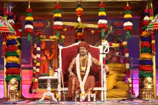A seated man in an orange-brown ochre robe and matching turban sits on an ornate red-cushioned white throne-style chair on a stage decorated with multicoloured tassel and flower garlands in red, yellow, blue, green and white. He wears a pink and white flower garland, gold necklaces, a gold bracelet and a microphone is in front of him. Behind him is a deity murti with multiple arms, and golden lion figures flank the stage. Golden cushions are piled on the right and a small side table holds a glass.