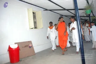 A photograph of a covered walkway beside a whitewashed wall with a barred window and a red bin on the left. A man with long dark hair wears a saffron-orange robe and sandals and walks forward, with one hand raised near his shoulder. Several men in white dhotis and white shirts, some with orange shawls, follow or walk alongside him, and a hanging potted plant hangs from a blue pole on the right. The probable Swamiji is the man in the orange robe with long swept-back hair, shown here in the group procession.