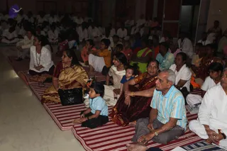 A large seated audience sits on striped red-and-white mats in a hall during a discourse or satsang. In the foreground, a woman in a gold-and-maroon sari sits beside an older man in a light blue striped shirt and grey trousers, and a young boy in a blue shirt sits cross-legged nearby. Behind them, rows of devotees wear white and orange clothing, and several women wear saris in green, maroon and blue. The crowd faces forward toward the speaker, with a mix of families and children.