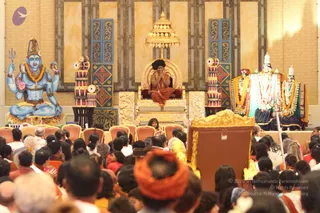 A large hall with a crowd of seated devotees facing a golden throne-like seat on a raised stage. On the throne sits a figure with long dark swept-back hair, wearing an orange-saffron robe and a garland of beads, hands pressed together in greeting. Behind him is a large painted blue deity figure with four arms, a trident and a tiger skin, and to the right stand two white-and-gold deity murti decorated with garlands. Ornate panels, drums and a gilded canopy surround the stage. In the foreground, a person in an orange head wrap sits with their back to the camera, and a small child is visible at the front of the crowd.