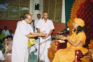 A photograph of a seated figure on an ornate carved wooden throne with red velvet upholstery, wearing an orange-saffron top and matching orange head wrap, with a long dark braid and a rudraksha-style mala. A man in white dhoti and shirt stands at the left with hands extended in front of him, another man in white shirt stands behind a microphone on a stand, and gold items sit on a tray near the throne. Seated devotees in white and pink saris, one woman wearing a white flower garland, and other men in white clothing fill the background of a hall with lattice windows.