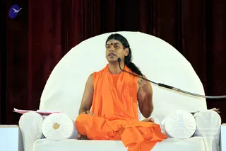 A seated man in orange saffron robes speaks into a gooseneck microphone on a white cushioned throne with round bolster pillows. He has long dark hair pulled back, a red bindi on his forehead, and wears a saffron cloth draped over his left shoulder, leaving the right arm bare. His hands rest in his lap, and papers sit on the left armrest. Behind him hang deep maroon velvet curtains, with a small blue-and-white logo in the upper left corner. The appearance is consistent with a probable Swamiji (probable) based on the long hair, saffron clothing and rudraksha-style adornment cues, shown seated in discourse on the throne.