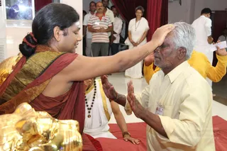 A seated man in a maroon and gold-bordered shawl places his right hand on the forehead of a kneeling older man with grey hair and a short beard, who wears a cream shirt and trousers and an ID badge on his chest with his palms pressed together in reverence. The seated figure has long dark hair tied up with a red band, a rudraksha mala, and a gold necklace, which are the cues for a probable Swamiji in an in_person blessing scene. Behind them a seated attendant in white with a rudraksha mala and a gold-bordered cloth watches, and onlookers stand near a red curtain in the background, including a man in a striped polo shirt and a woman in white. A gold ornament sits in the lower left corner, and a red cloth covers the floor. The scene shows a forehead blessing given to a devotee.