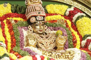 Close-up photograph of a gilded deity murti decorated with dense garlands of yellow marigolds, red roses and green leaves. The figure wears a tall white-and-red beaded crown with a central ornament, a heavily jewelled gold necklace set with red stones, and raised gold hands, one holding a green fruit. Its face is gold-plated with painted eyes. Fresh flower decoration frames the murti against a gold-edged background. No people are visible.