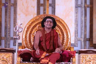 A man seated cross-legged on an ornate gold throne with a carved circular backrest, set against a cream and gold panelled backdrop. He wears a deep red robe with an orange-gold edge, a thick red rudraksha mala with a matching double strand, a gold necklace, and a white tilak with a red dot on his forehead. His hair is long and dark, and he has a bracelet on his wrist and bare feet. A silver trident stands to the left and a leopard-print cushion rests on the seat. Probable Swamiji, seated on the throne, with appearance cues of long hair, red robes, rudraksha beads and gold necklace. Only one physical person is visible.