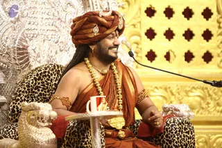 A smiling man seated on an ornate silver throne upholstered in leopard-print fabric, in front of a gold carved panel with a lattice of cut-out diamond shapes. He wears a rust-orange turban with a white crescent-shaped ornament and a jewelled brooch, a saffron-orange shawl over bare shoulders, long gold beaded necklaces, a rudraksha mala, gold armlets and a gold waist ornament. He gestures with his right hand while a small white cup sits on a silver side table beside him, and a microphone on a stand is at right. Probable Swamiji (probable) seated on the throne, identified by the saffron/orange clothing, ornate gold necklaces, rudraksha mala and turban cues.
