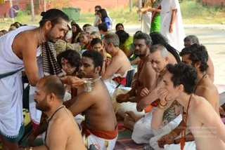 Outdoor photograph of a seated group of men, mostly bare-chested in white dhoti-style cloth with orange or red waist cloths, many wearing rudraksha malas and forehead markings. On the left, a standing man with hair tied up and a horizontal tilaka wears a white dhoti and checked shawl and pours liquid from a small metal cup into the cupped hands of a seated devotee. Other participants sit on striped mats on the ground, some with hands folded, while women in saris and onlookers sit or stand in the background near a red brick wall. The scene resembles a ritual or puja gathering. No clearly identifiable a person figure is evident from appearance, so the attendees are described by role.
