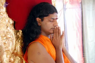 A man with long, dark, swept-back hair and a red bindi on his forehead sits in profile with his eyes closed and palms pressed together in namaskar. He wears a sleeveless saffron/orange garment. He leans against a red cushion beside an ornate gold carved throne-like frame, with a white curtain behind him.