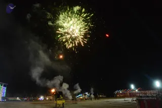 Night scene at an outdoor ground with a large burst of green and gold fireworks lighting the sky above. Smoke drifts from a fireworks launcher and cart in the foreground, with a bright orange flare near the base. Strings of coloured lights outline a stage structure at left, and a red tent-like pavilion with lit poles stands at right. A few small figures and parked white vehicles are visible in the distance across the open ground.