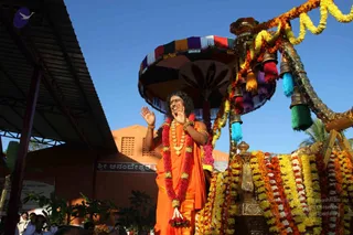 Outdoor festival procession scene under a clear blue sky. A man in a saffron-orange robe with long swept-back dark hair stands on an ornate golden palanquin decorated with marigold and red garlands, orange and yellow flower strands, and a multicolored tasselled canopy in red, purple, teal and green. He wears a red and white flower garland, a gold necklace and gold bangles, and raises both hands in an expressive gesture. Behind him is an orange-painted building with a sign reading Sri Anandeshwara, and a few onlookers stand at the lower edge of the frame.