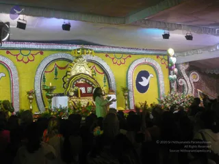 A photograph of a large event stage decorated in yellow with white and red arches, floral borders and a dove emblem on a navy roundel. In the centre, a golden ornate throne-like seat with a red cushion sits beneath a canopy of yellow and white garlands, flanked by brass oil lamps and flower arrangements. A woman in a light green salwar kameez stands at the front of the stage, one arm raised and gesturing toward a seated area. A dense crowd of devotees, mostly seen from behind, fills the foreground, with many raising hands. Overhead, stage lights hang from white drapery.