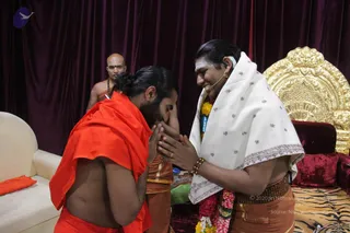 A photograph of a devotee in an orange saffron cloth bowing deeply with palms pressed together, touching his forehead to the joined hands of a man in a white shawl with red embroidered motifs. The standing man wears a small microphone headset, a red and white flower garland, a brown beaded waist belt and orange dhoti, and smiles warmly. Behind them stands a bare-chested man with a rudraksha mala and orange cloth. Behind the seated figure is an ornate gold throne on a tiger-striped cushion, with burgundy velvet drapes and a white sofa with an orange cushion on the left. The scene suggests a respectful greeting or seeking blessing.