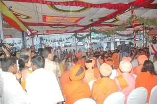 A large crowd gathers under a white and red festival canopy decorated with patterned yellow, green and red bands and hanging green leaves. Many people wear saffron-orange robes and turbans, while others wear white kurtas. In the centre a man with long hair and a saffron robe stands among seated sadhus in orange and pink turbans. Video cameras are raised on the left, and white chairs are arranged in the foreground. Behind the canopy, a poster-sized portrait and white-clad attendants are visible beyond a red-and-white striped pole.