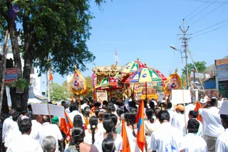 A street procession in a small Tamil Nadu town on a bright day. A decorated ratha or palanquin with a golden canopy, gold ornaments and a deity murti is pulled along the road under colourful striped parasols with gold finials. Crowds of devotees in white clothes walk alongside, many holding saffron and orange flags, and several carry blank white placards on poles. Women in cream and yellow saris with long braids and flowers in their hair walk in the crowd. Utility poles, a tree and shopfronts line the roadside.