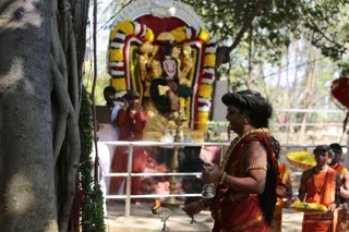 Outdoor photograph of a ritual at a shrine beneath a large banyan tree. A figure with long swept-back dark hair and gold necklaces, wearing a red-orange garment with a gold-edged shawl and gold armlet, stands holding a small metal bell-like vessel in one hand and a lit oil lamp on a tray in the other. Behind a metal railing, a golden deity murti is framed by garlands of yellow marigolds and colourful striped flower strings. Two men in orange and saffron clothing stand to the right, one holding a basket of yellow flowers, and a man in red near the shrine raises his hands in prayer. Green leaf garlands hang on the tree trunk.